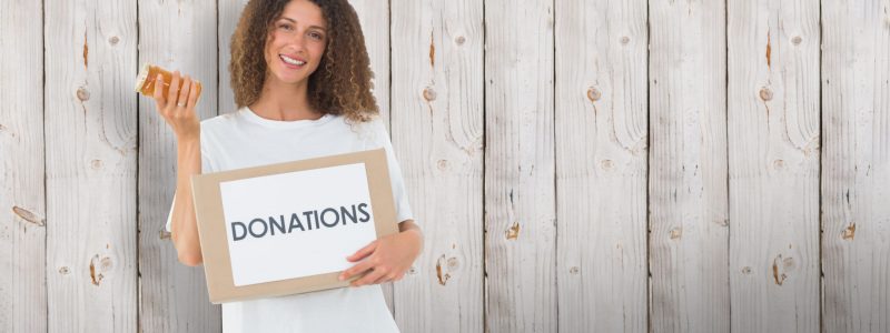 Happy volunteer holding a box of donations and jam jar against wooden background